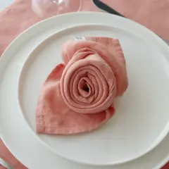 Pliage serviette rose en forme de rose sur une assiette blanche, avec une fourchette à gauche et des raisins à côté.
