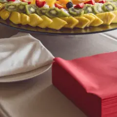Une table de restaurant prête pour un repas, avec un plateau de fruits frais, des couverts emballés dans des serviettes blanches et des serviettes rouges pliées.