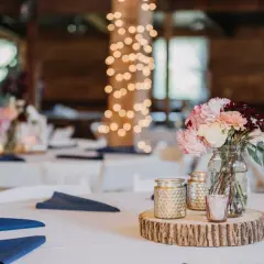 Un bouquet de fleurs roses et crème sur une tranche de bois, entouré de bougies dorées. Le **protocole fiançailles** est respecté avec des serviettes bleues sur des tables blanches, sous des guirlandes lumineuses.