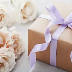Un cadeau d'anniversaire fait maison, joliment emballé dans du papier kraft et un ruban lilas, entouré de roses pâles.