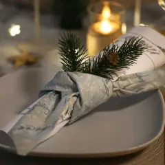 Une serviette en tissu, décorée d'une branche de sapin et d'une pomme de pin, est pliée en forme de smoking sur une assiette.