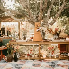 Un magnifique buffet de mariage thème automne, avec un gâteau orange, des fleurs séchées et des pampas sous un grand arbre.