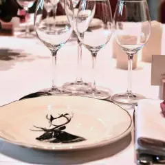 Une table dressée pour un dîner de gala, avec une assiette ornée d'un cerf, des verres à vin et une carte de visite "S.A.R. LE Grand-Duc de Luxembourg". La fourchette à la française est prête pour le festin.