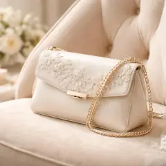 Pochette mariage champêtre en satin ivoire, ornée de dentelle florale et d'une chaîne dorée, posée sur un fauteuil crème.