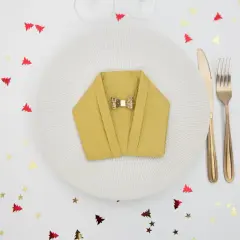 Pliage serviette nouvel an en forme de veste, orné d'un nœud papillon doré, sur une assiette blanche. Confettis sapins et étoiles rouges et dorés.