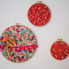 Décoration murale avec trois cercles à broder. Un grand cercle fleuri sert de rangement pour des ciseaux et un stylo, orné d'un ruban à pompons. Deux autres cercles rouges décoratifs complètent l'ensemble.