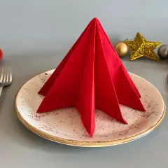 Un sapin en serviette papier rouge décore une assiette, prêt pour un repas festif. Des boules de Noël et une étoile dorée complètent la mise en scène.