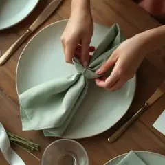 Mains créant un pliage serviette simple et beau, vert pâle, sur une assiette blanche. Une table dressée pour un repas élégant.