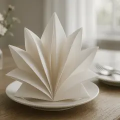 Une serviette en papier pliée en accordéon sur une assiette, prête pour un repas. Un bouquet de fleurs est visible en arrière-plan.