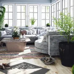 Un salon lumineux et aéré, baigné de lumière naturelle, avec un canapé d'angle confortable, des plantes luxuriantes et une décoration au thème bucolique.