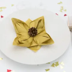 Une serviette dorée pliée en forme de fleur de lotus, ornée d'une pomme de pin, sur une assiette blanche. Confettis sapins rouges et dorés sur la table.