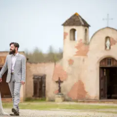 Un couple rayonnant, tout juste marié, se promène main dans la main devant une chapelle rustique, évoquant un mariage western.