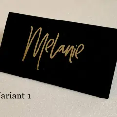 Marque place noir et or avec le prénom "Melanie" écrit en calligraphie dorée. Un élégant détail pour une table de fête.