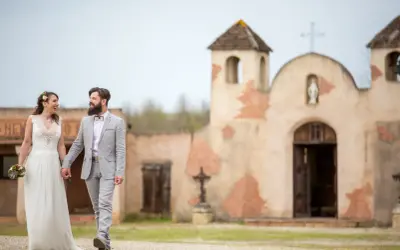 Un couple rayonnant, tout juste marié, se promène main dans la main devant une chapelle rustique, évoquant un mariage western.