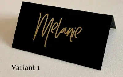 Marque place noir et or avec le prénom "Melanie" écrit en calligraphie dorée. Un élégant détail pour une table de fête.