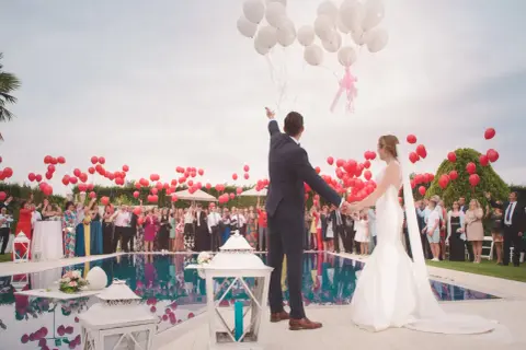 Un couple de mariés, entouré d'invités, lâche des ballons blancs et rouges. Un lâcher de ballon mariage symbolisant l'amour et l'espoir.