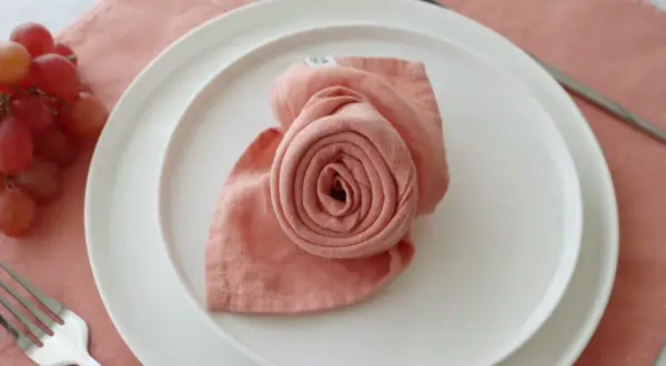 Pliage serviette rose en forme de rose sur une assiette blanche, avec une fourchette à gauche et des raisins à côté.