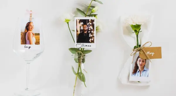 Marque place fleur : photos de invités (Emy, Simon, Mary) attachées à des fleurs et des verres, pour une décoration de table personnalisée.