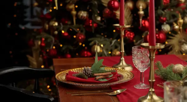 Une élégante deco table noel rouge et or, avec des bougies, des ornements dorés et un sapin scintillant en arrière-plan.