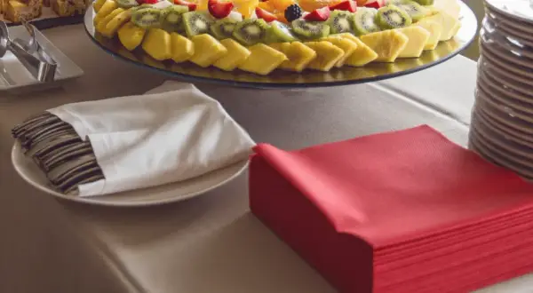 Une table de restaurant prête pour un repas, avec un plateau de fruits frais, des couverts emballés dans des serviettes blanches et des serviettes rouges pliées.
