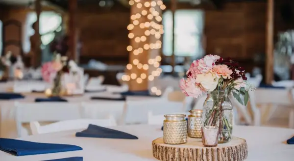 Un bouquet de fleurs roses et crème sur une tranche de bois, entouré de bougies dorées. Le **protocole fiançailles** est respecté avec des serviettes bleues sur des tables blanches, sous des guirlandes lumineuses.