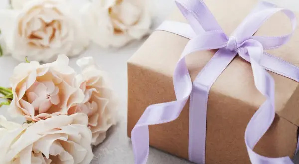 Un cadeau d'anniversaire fait maison, joliment emballé dans du papier kraft et un ruban lilas, entouré de roses pâles.