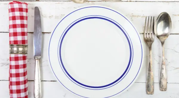 Une assiette blanche à double bordure bleue, une serviette à carreaux rouges et blancs, un couteau, une fourchette et deux cuillères. Qui a inventé les couverts ?