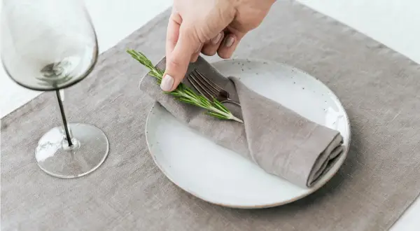 Une main dépose une branche de romarin sur un pliage de serviette mariage boheme, sur une assiette blanche.