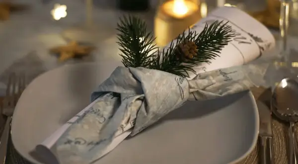 Une serviette en tissu, décorée d'une branche de sapin et d'une pomme de pin, est pliée en forme de smoking sur une assiette.