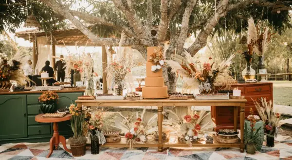 Un magnifique buffet de mariage thème automne, avec un gâteau orange, des fleurs séchées et des pampas sous un grand arbre.