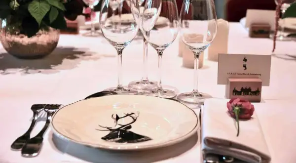 Une table dressée pour un dîner de gala, avec une assiette ornée d'un cerf, des verres à vin et une carte de visite "S.A.R. LE Grand-Duc de Luxembourg". La fourchette à la française est prête pour le festin.