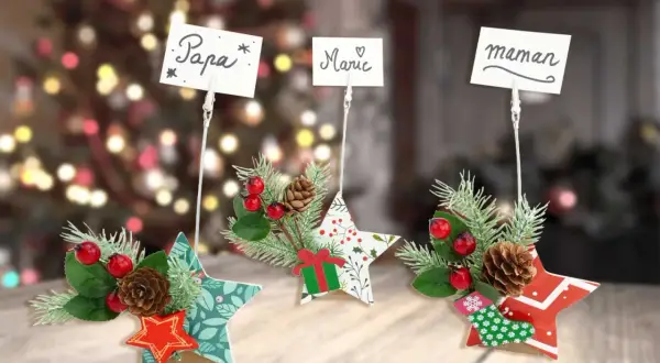 Porte nom original à faire soi-même : étoiles décorées de branches de sapin, baies rouges et pommes de pin, avec des noms écrits sur des cartes.