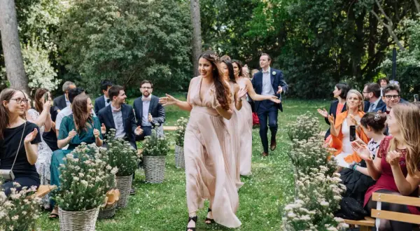 Le cortège d'église pour ce mariage en extérieur est joyeux. Les demoiselles d'honneur, en robes fluides, marchent sur l'herbe, applaudies par les invités.