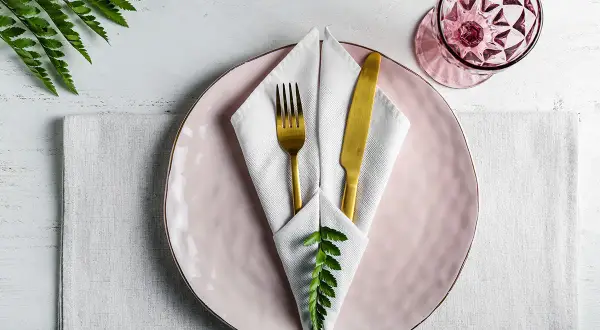 Service de table élégant avec pliage serviette communion, assiette rose, couverts dorés et verre à facettes.