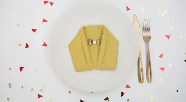 Pliage serviette nouvel an en forme de veste, orné d'un nœud papillon doré, sur une assiette blanche. Confettis sapins et étoiles rouges et dorés.