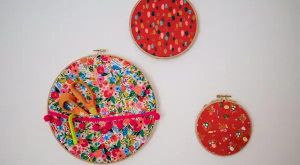 Décoration murale avec trois cercles à broder. Un grand cercle fleuri sert de rangement pour des ciseaux et un stylo, orné d'un ruban à pompons. Deux autres cercles rouges décoratifs complètent l'ensemble.