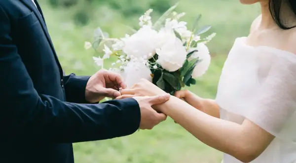 Un homme en costume met une bague à la main d'une femme, bouquet de fleurs blanches. Un poème mariage Baudelaire pour un amour éternel.