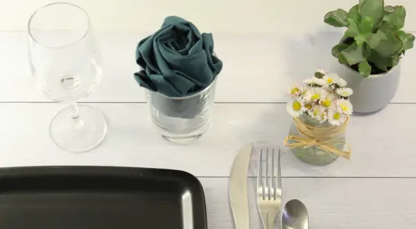 Un verre à pied, une serviette pliée en rose dans un verre, des petites fleurs et des couverts sur une table en bois.