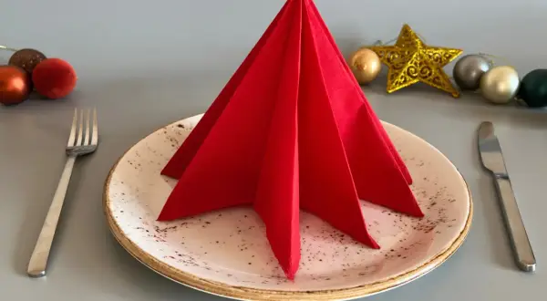 Un sapin en serviette papier rouge décore une assiette, prêt pour un repas festif. Des boules de Noël et une étoile dorée complètent la mise en scène.
