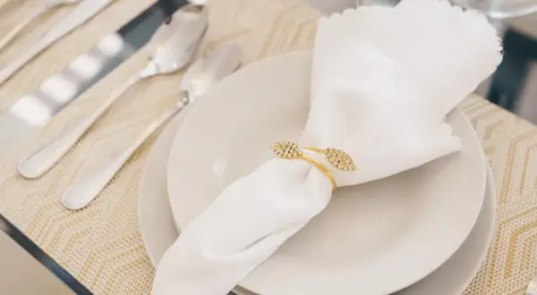 Une serviette blanche mariage, ornée d'un anneau doré scintillant, repose sur une assiette. Des couverts argentés sont disposés à côté.