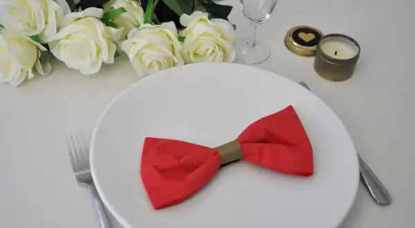 Une assiette blanche ornée d'un pliage serviette noeud papillon rouge. Des roses blanches et une bougie complètent la décoration.