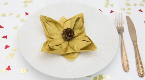 Une serviette dorée pliée en forme de fleur de lotus, ornée d'une pomme de pin, sur une assiette blanche. Confettis sapins rouges et dorés sur la table.