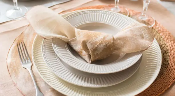 Une table dressée avec des assiettes empilées, une serviette en tissu nouée, une fourchette et des verres. La différence entre faïence et porcelaine est subtile, mais la qualité est évidente.