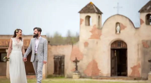 Un couple rayonnant, tout juste marié, se promène main dans la main devant une chapelle rustique, évoquant un mariage western.