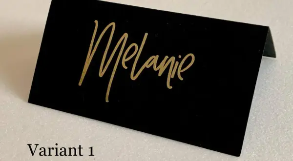 Marque place noir et or avec le prénom "Melanie" écrit en calligraphie dorée. Un élégant détail pour une table de fête.