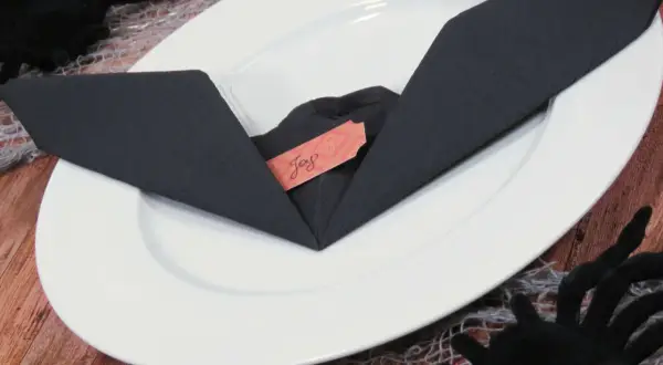 Une serviette noire pliée en forme de chauve-souris sur une assiette blanche, avec une étiquette "Joy". Décoration d'Halloween.