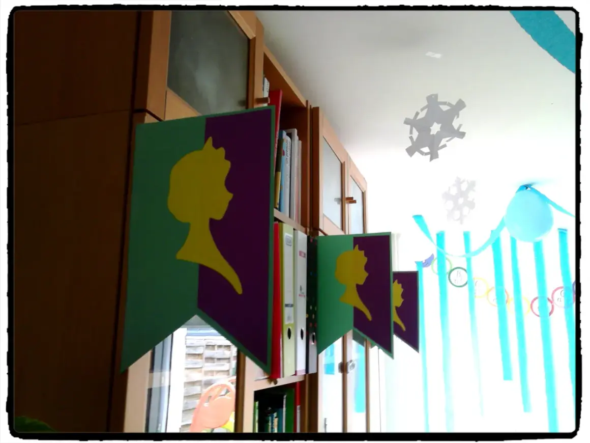 Guirlande "décoration reine des neiges à faire soi même" avec silhouettes jaunes sur fond violet et vert, suspendue devant une bibliothèque.