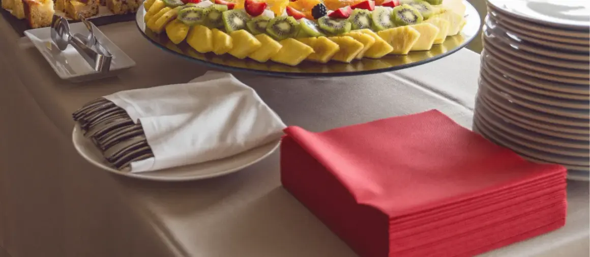 Une table de restaurant prête pour un repas, avec un plateau de fruits frais, des couverts emballés dans des serviettes blanches et des serviettes rouges pliées.