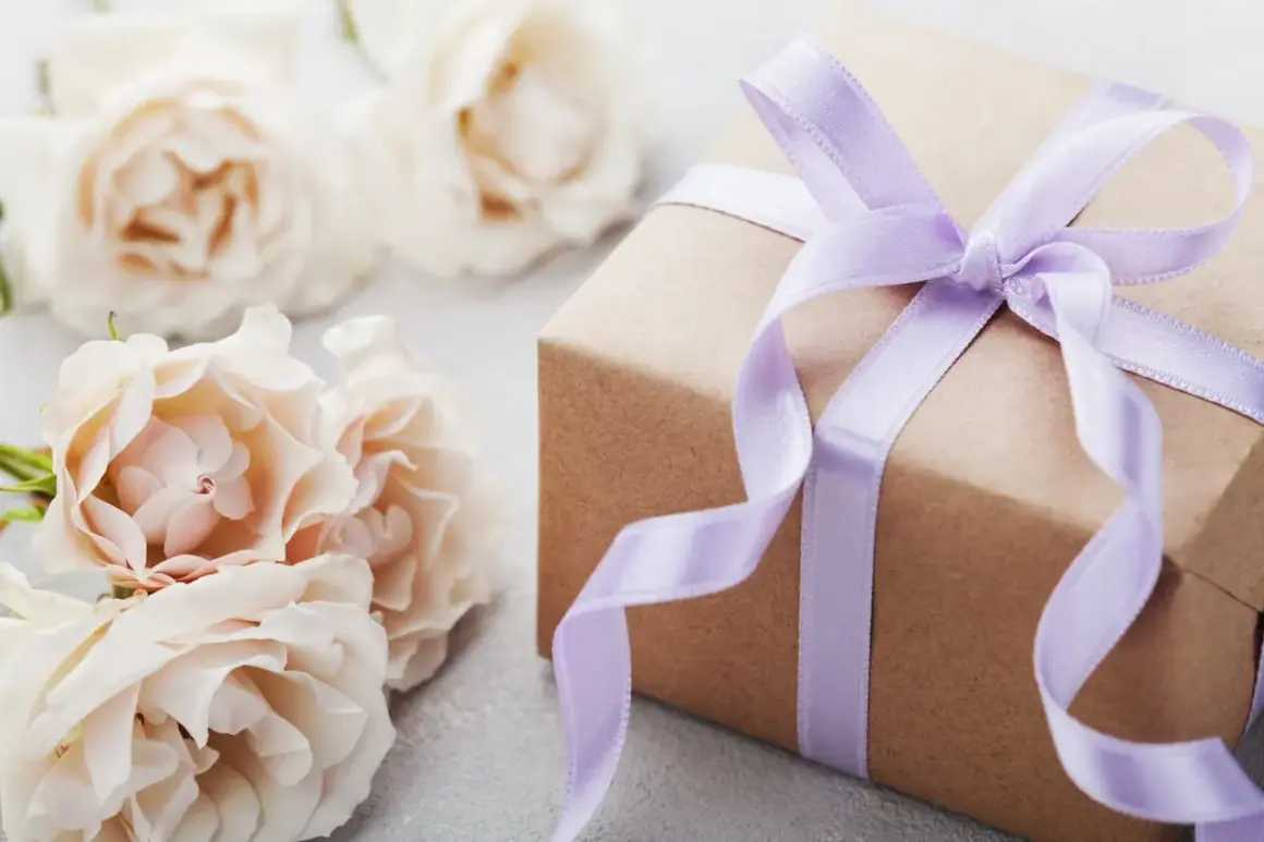 Un cadeau d'anniversaire fait maison, joliment emballé dans du papier kraft et un ruban lilas, entouré de roses pâles.