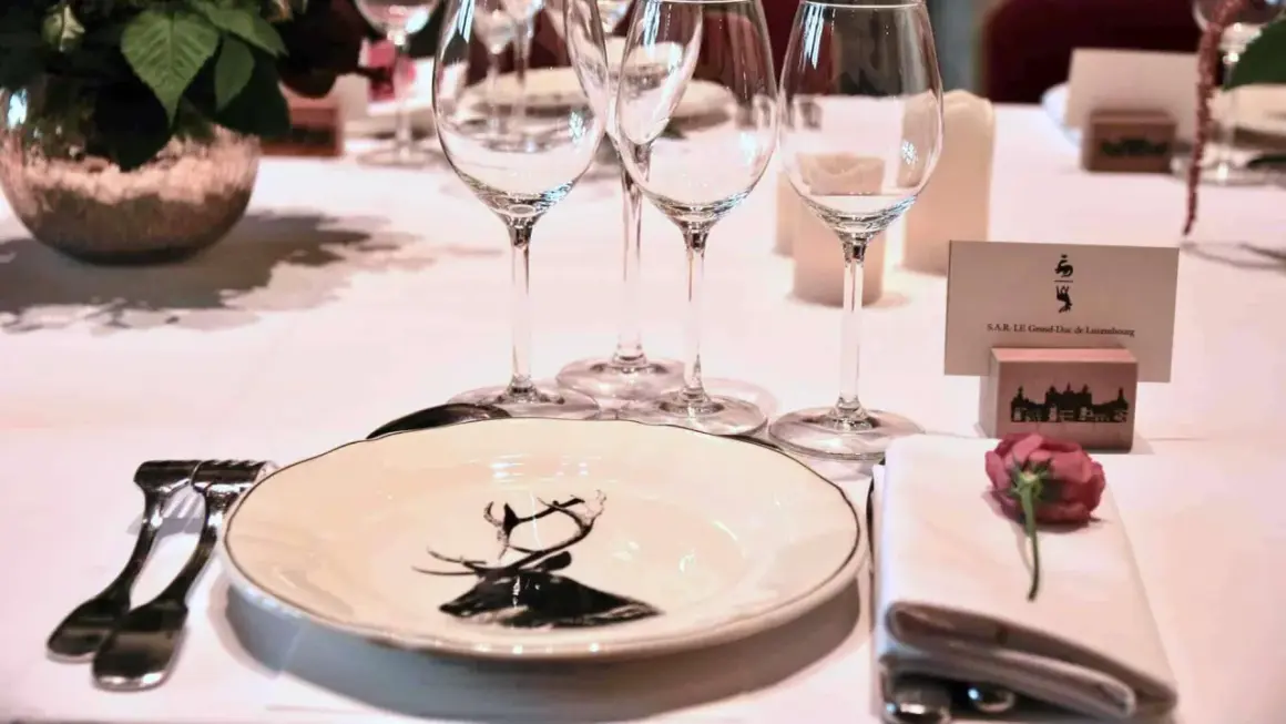Une table dressée pour un dîner de gala, avec une assiette ornée d'un cerf, des verres à vin et une carte de visite "S.A.R. LE Grand-Duc de Luxembourg". La fourchette à la française est prête pour le festin.